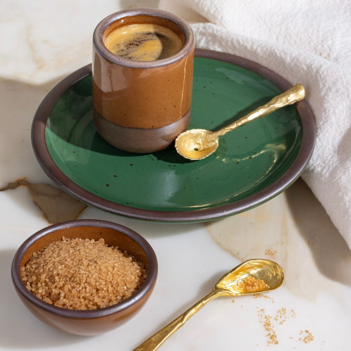 Glossy brown ceramic espresso cup on a green plate with brass shell spoon, next to a bowl of raw sugar on marble.