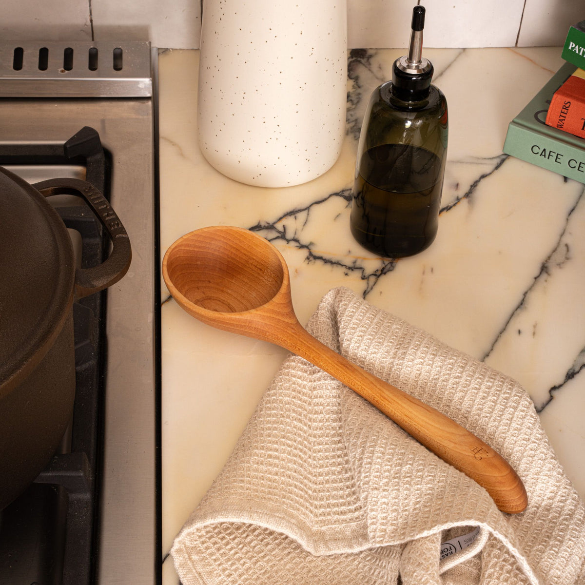 A wooden ladle with a large scoop next to a stovetop with a waffle weave kitchen towel and cruet.