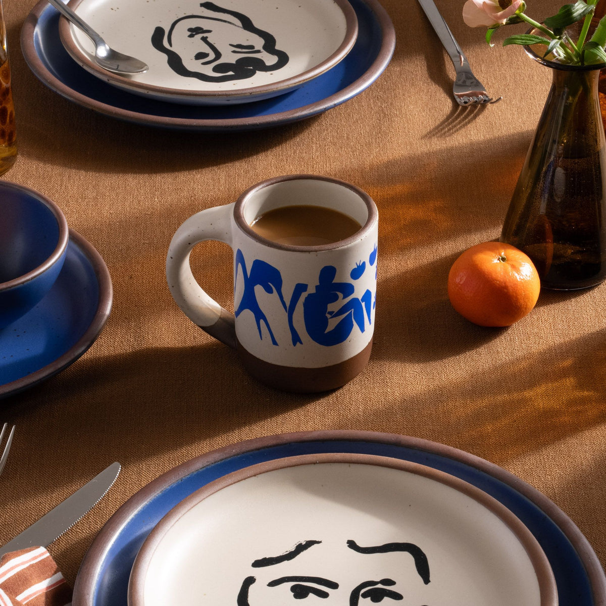 On a table, sits East Fork’s The Mug in a warm off white color with handle with a blue decal of Henri Matisse’s Femmes et singes, and a unglazed rim and bottom base, sits on a table with more ceramic dinnerware.