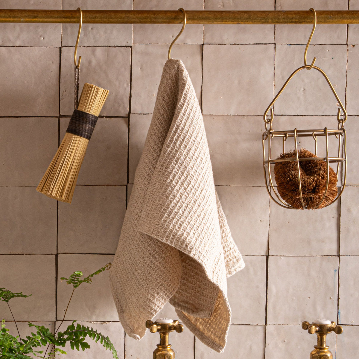 A natural waffle weave kitchen towel, scrub brush, and straw scrub hanging on a brass rod in front of a tiled wall above a sink.
