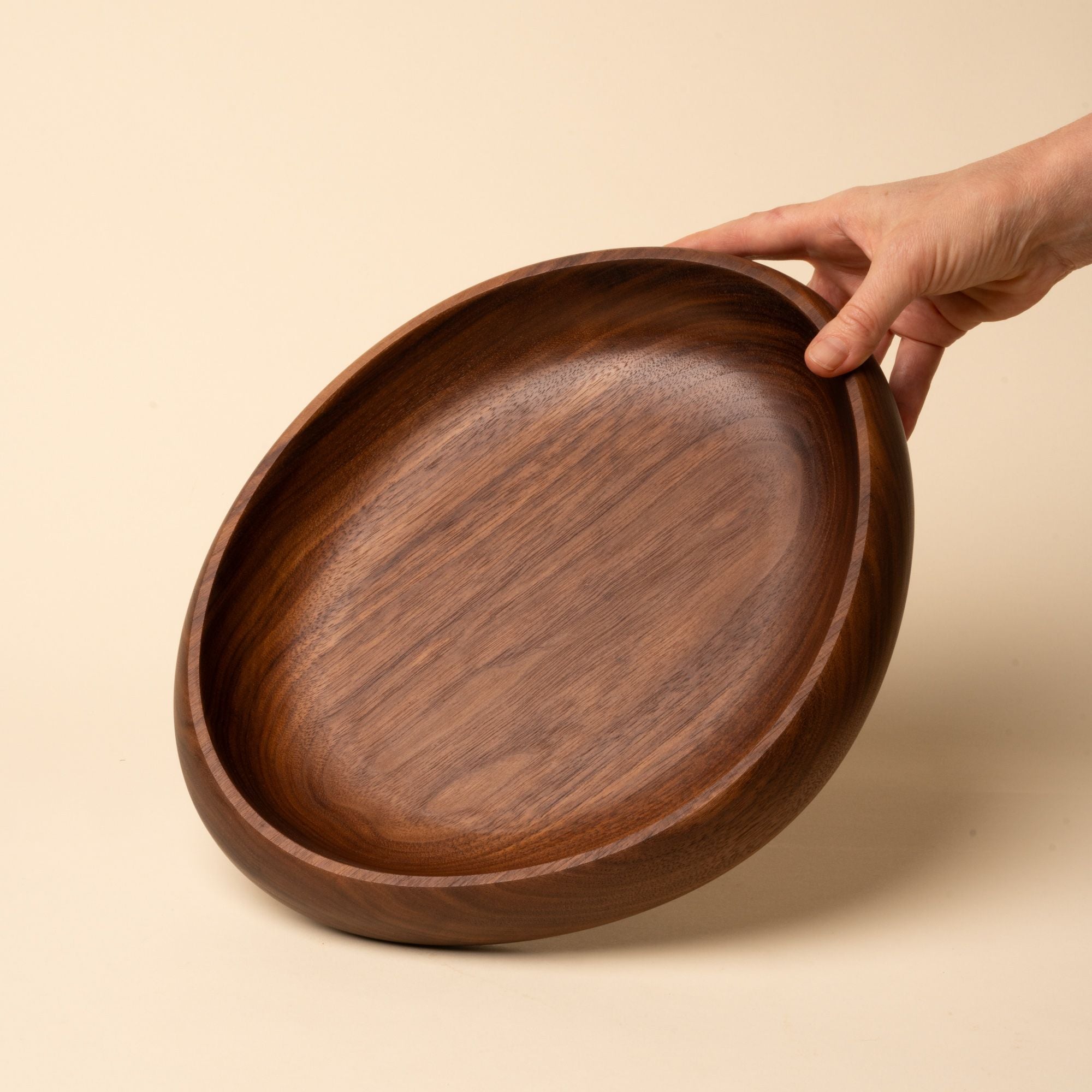 Hand holds out a shallow carved round walnut wood dish with smooth curved edges, resting on a beige background.