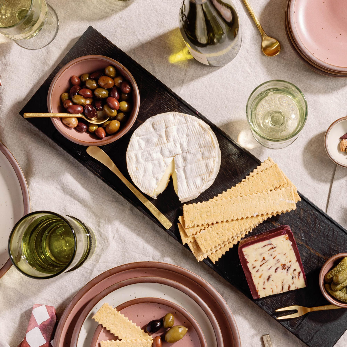 Cheese board with crackers, olives, and soft cheese on a dark platter, surrounded by pink dishes, glassware, and brass serving utensils.