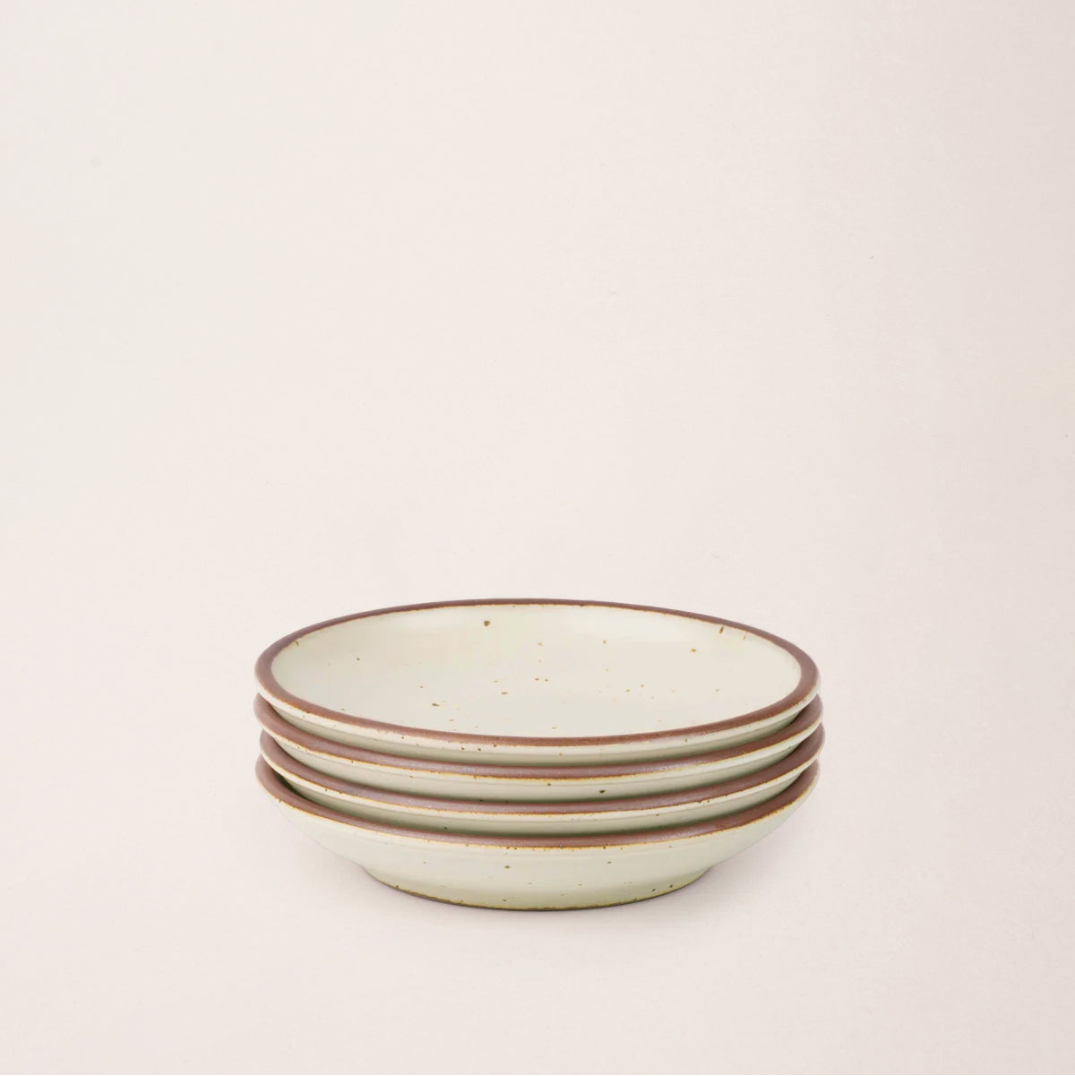 A stack of 4 small ceramic plate with a curved bowl edge in a warm, tan-toned, off-white color featuring iron speckles and an unglazed rim.