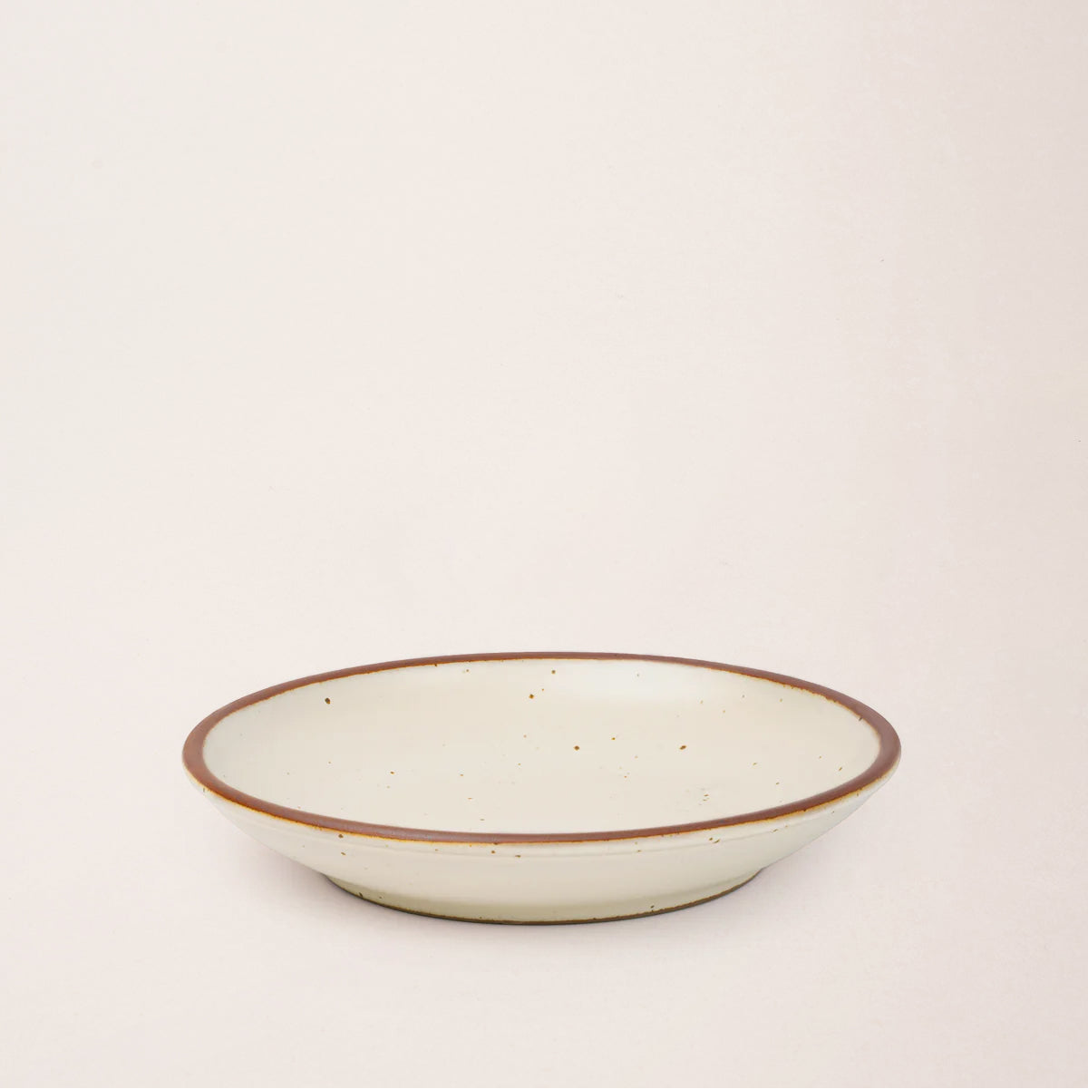 A medium side ceramic plate with a curved bowl edge in a warm off-white color featuring iron speckles and an unglazed rim.