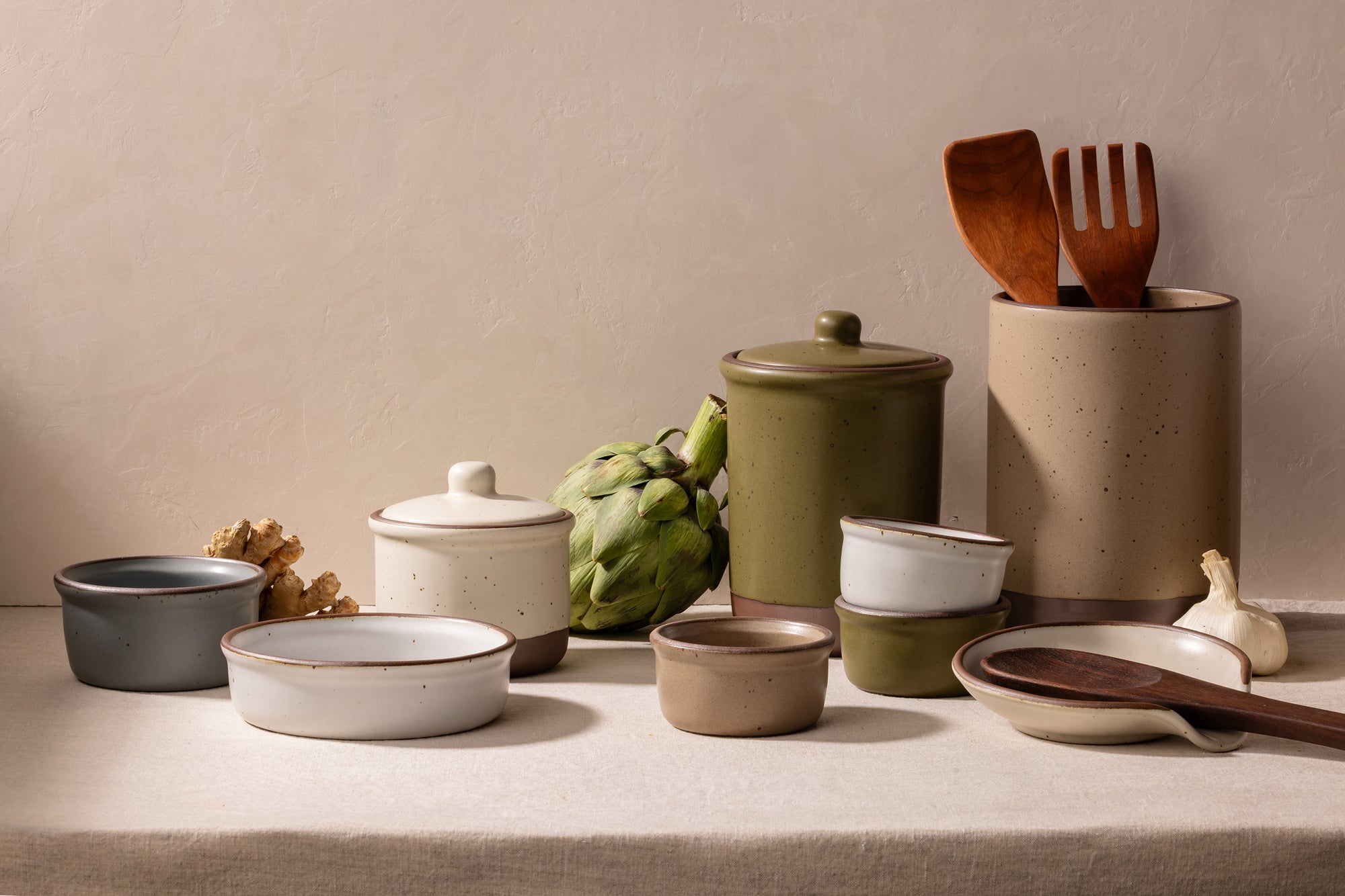 A styled collection of ceramic kitchenware on a linen-covered surface against a textured beige wall, including canisters, bowls, a lidded jar, wooden utensils, ginger, an artichoke, garlic, and a ceramic spoon rest.