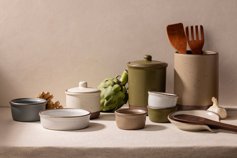 A styled collection of ceramic kitchenware on a linen-covered surface against a textured beige wall, including canisters, bowls, a lidded jar, wooden utensils, ginger, an artichoke, garlic, and a ceramic spoon rest.