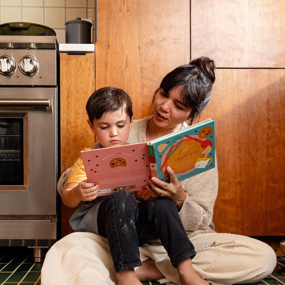 A parent and child sitting in the kitchen reading a children's illustrated cookbook about making cookies.