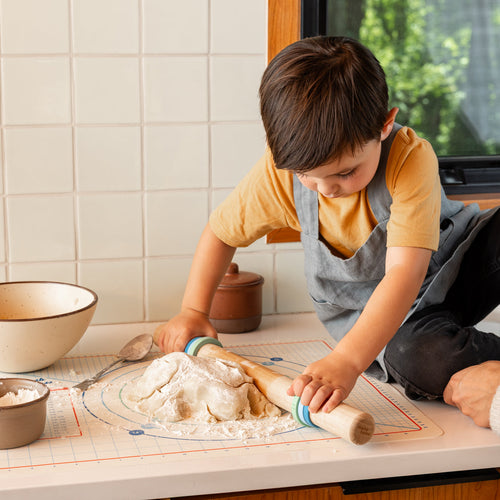 A child sitting on a kitchen counter rolling out dough with a rolling pin set and mat.