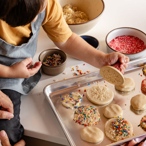A child holds a ceramic cookie snowflake stamp, pressing cookies on a baking sheet with sprinkles.
