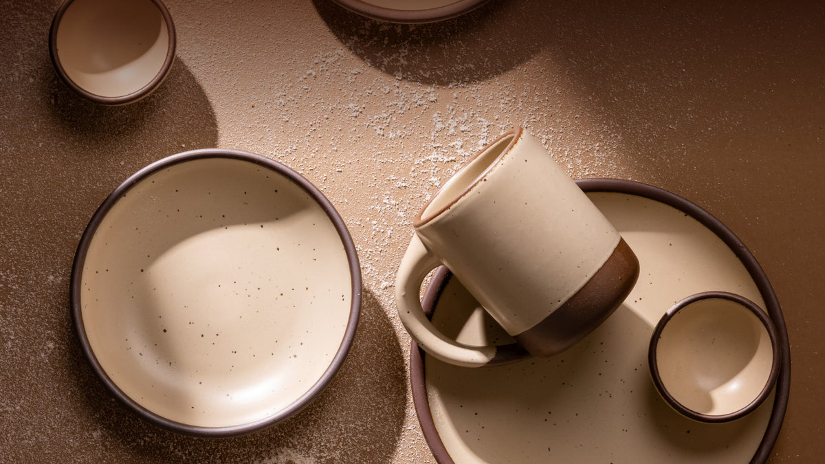 A set of handmade ceramic tableware in a warm cream color with unglazed base including a bowl, a mug resting on a plate, and a small bowl, arranged on a textured brown surface.