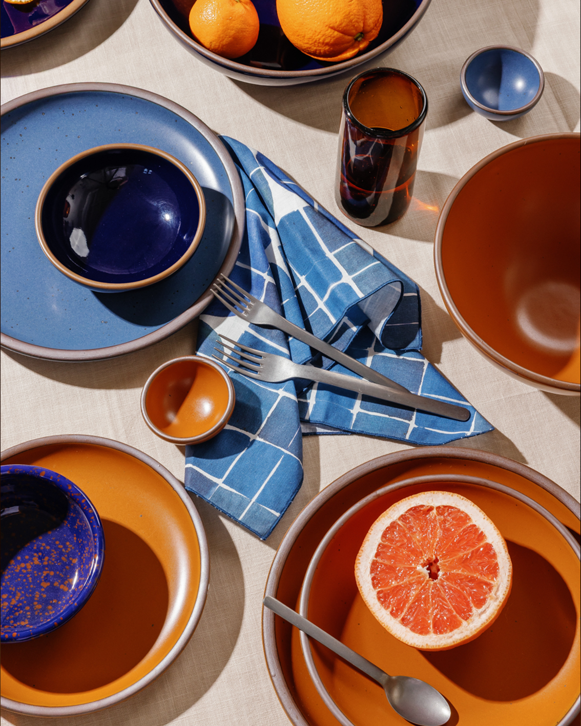 A styled tabletop dining scene with multiple ceramic plates and bowls in matte earthy orange and dark glossy blue, arranged with steel cutlery on a beige tablecloth. A blue-and-white check napkin, amber glass tumbler, oranges, and a halved grapefruit are also visible.