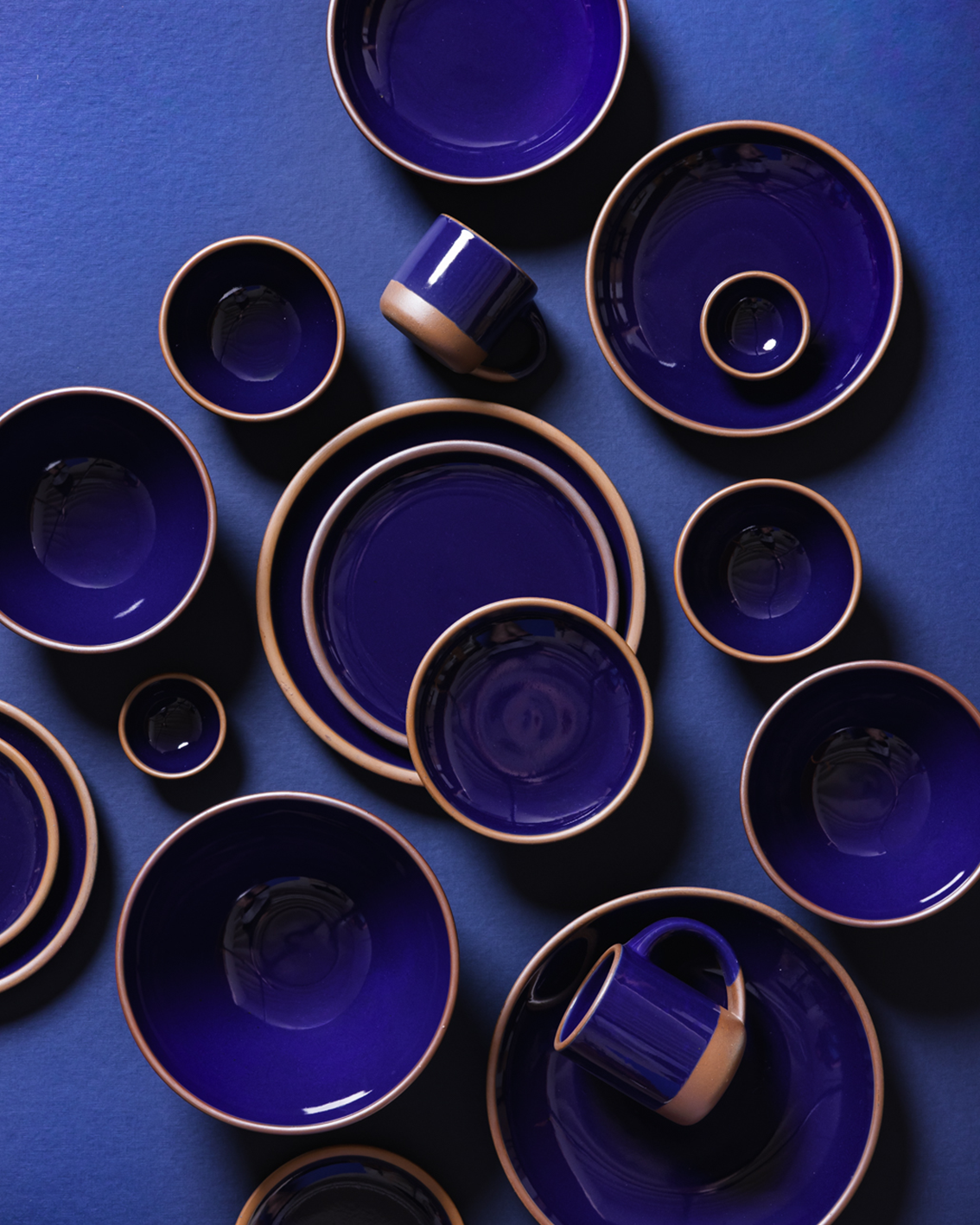A coordinated set of ceramic tableware in glossy dark blue with a contrasting light brown rim, arranged in a flat lay on a matching blue background. The set includes plates, bowls, cups, and mugs.