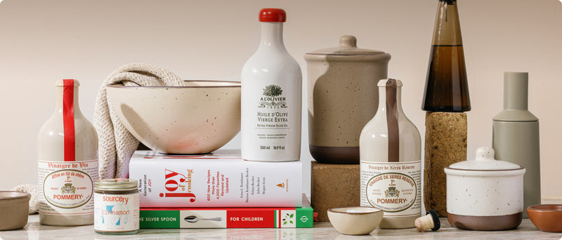 A styled product arrangement of kitchen tools and ingredients against a warm beige background, including ceramic bowls, bottles, jars, books, and a small towel.