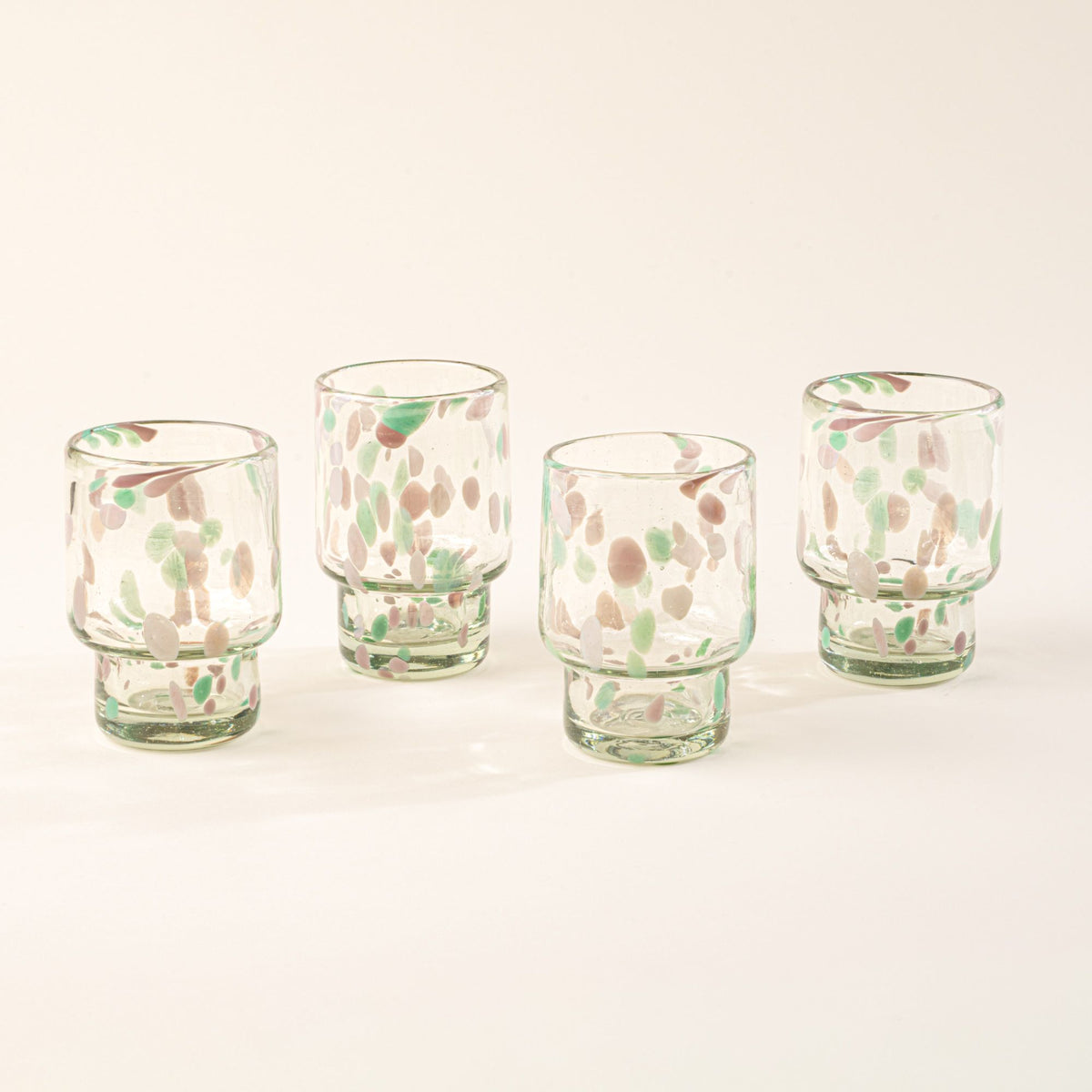 Four short clear glasses with pastel green and pink speckled patterns arranged in a row on a light background.