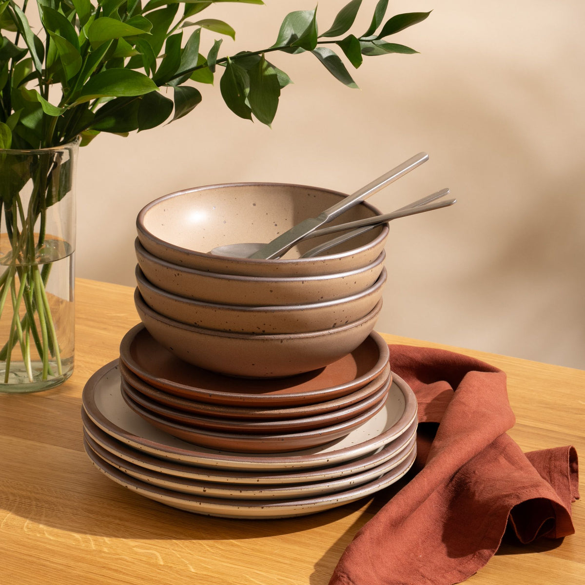 4 ceramic dinner plates in warm off-white, 4 side plates in cool terracotta, and 4 shallow dinner bowls in warm pale brown with iron speckles and unglazed rims, stacked on a table with silverware