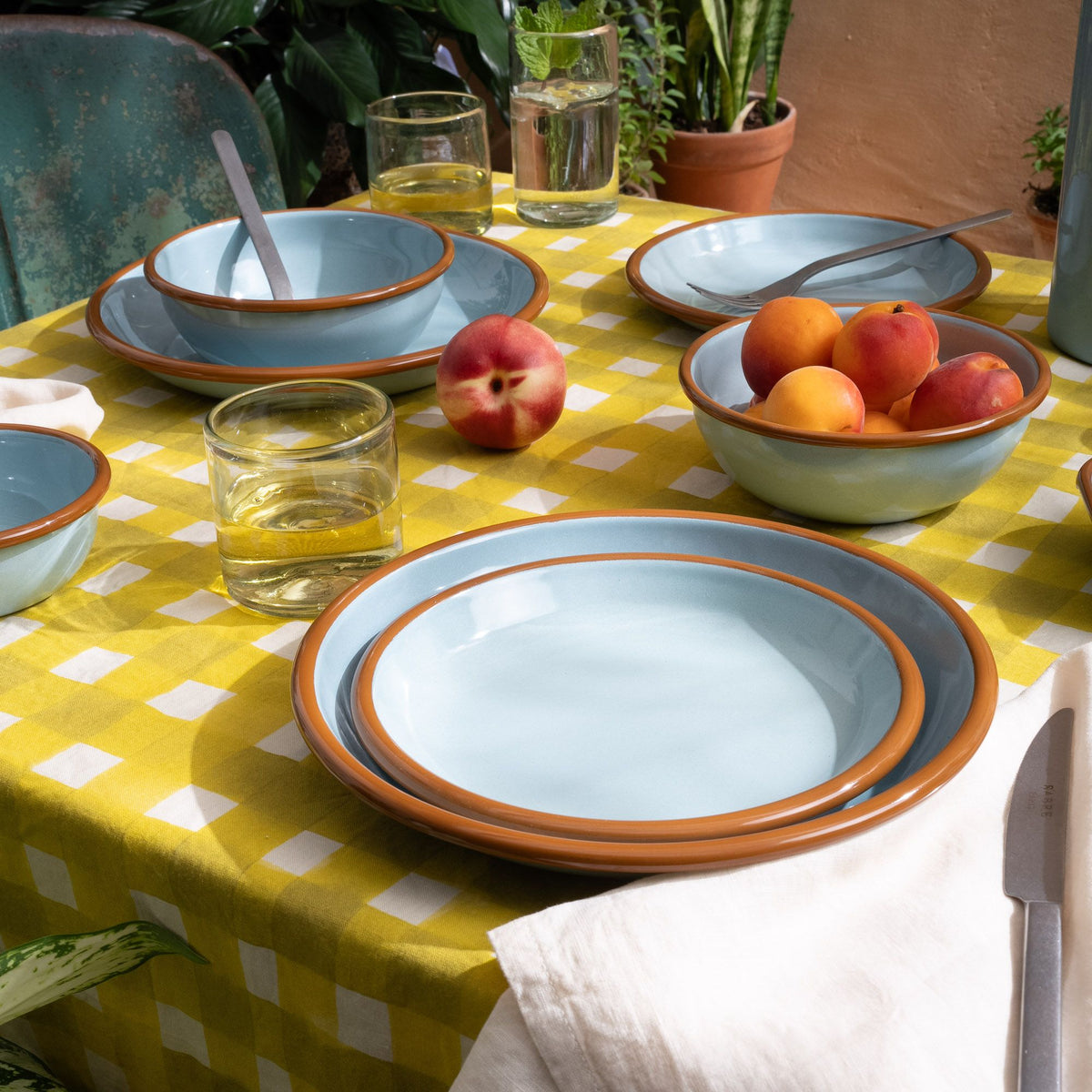 Short clear tumbler glasses on a table with robin's egg blue plates and bowls, on a yellow crosshatch tablecloth