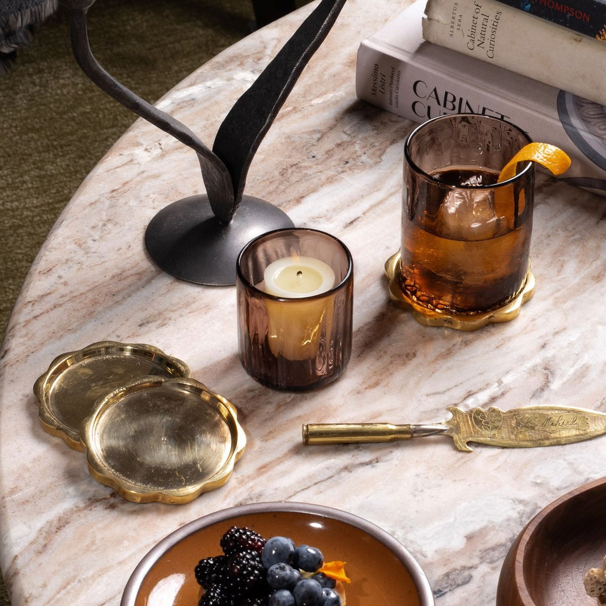 Brass scalloped coasters stacked on a marble table, one holding a glass of whiskey beside a candle and fruit plate.