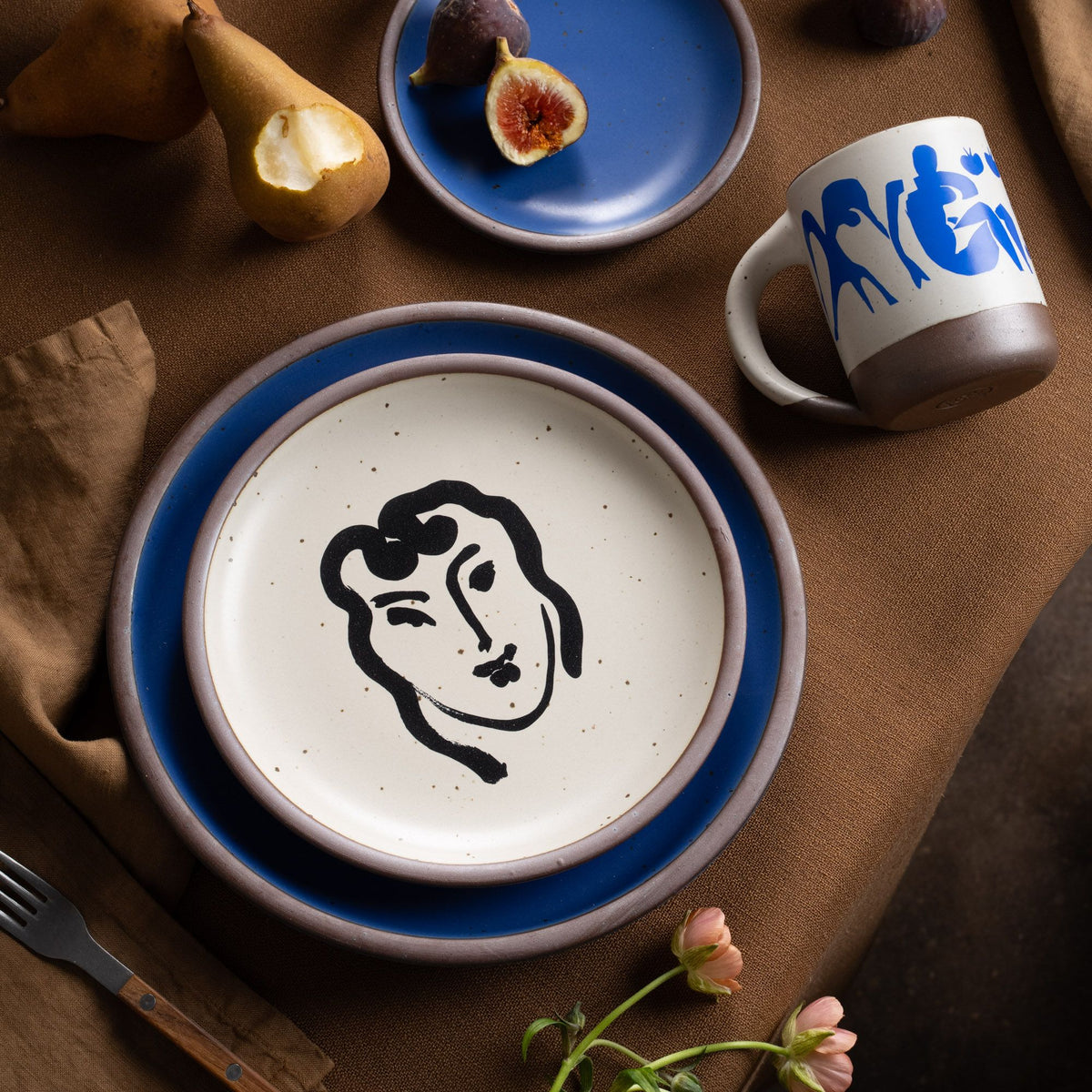 A ceramic plate featuring a decal of the artwork, Patitcha souriante, by Henri Matisse. A minimalist black ink decal of a woman's smiling face in an abstract, expressive style centered on an off-white ceramic plate with an unglazed rim, styled with a blue plate and other dishes on a table with flowers and fruit.