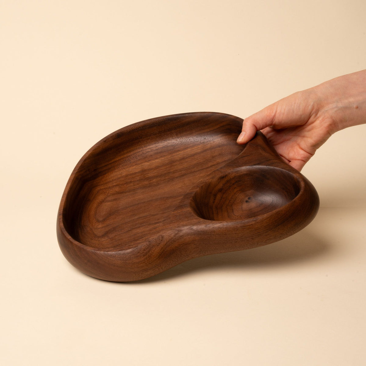 Hand holds out a carved walnut tray with smooth organic form, featuring two rounded compartments on a neutral backdrop.