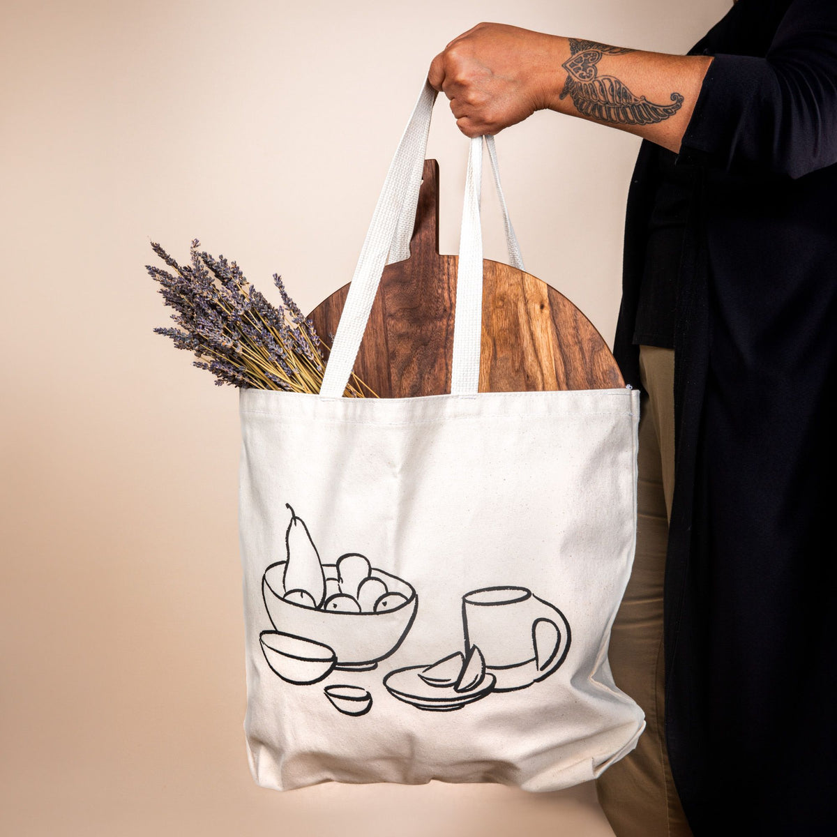 A cream canvas tote has a charcoal illustration on the front of a bowl with fruit, little bowls, plate, and a mug. A hand holds the tote and a large serving board and bundle of lavender is hanging out of the top.