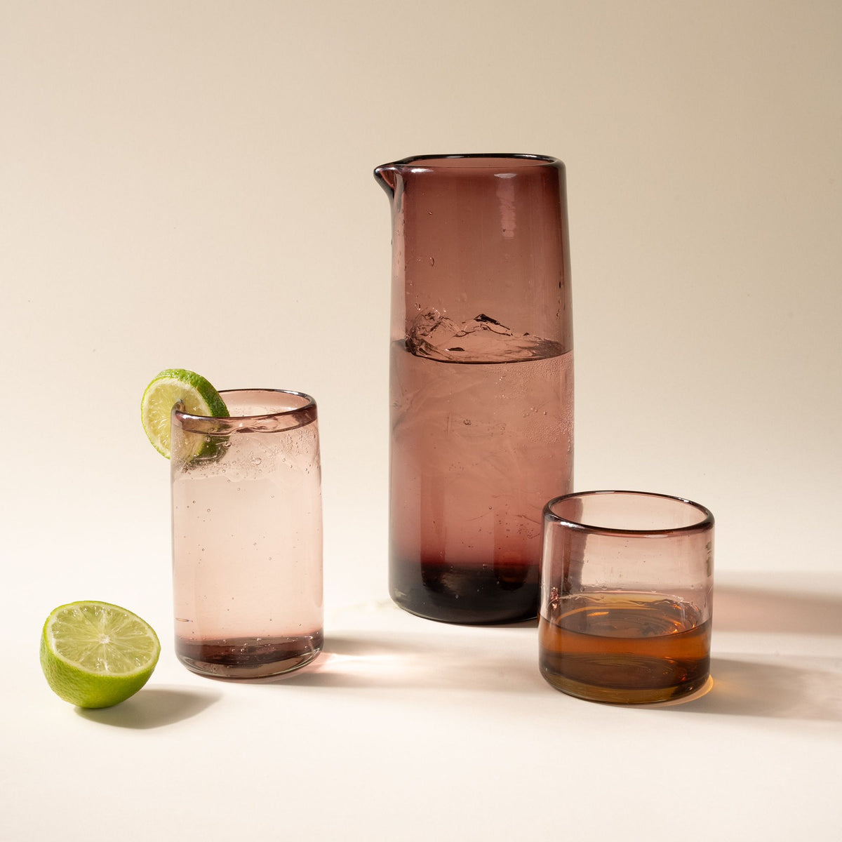 A tall tumbler, large pitcher, and short tumbler, of transparent glassware in a soft plum color, all filled with drinks.