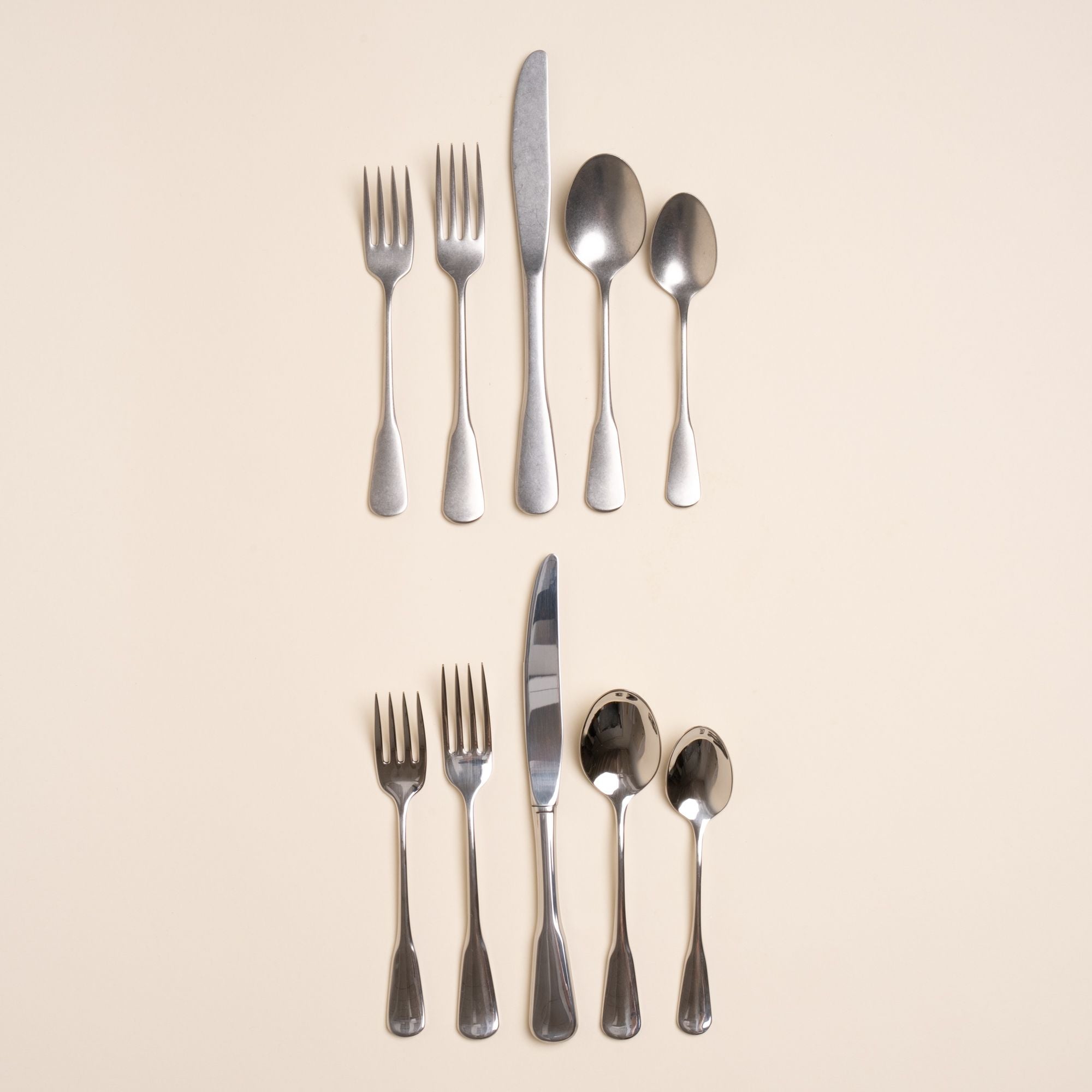 Two sets of stainless steel five-piece flatware including 2 forks, 2 spoons, and 1 knife. One is in a polished finish, the other in a tumbled matte finish, both a classic design