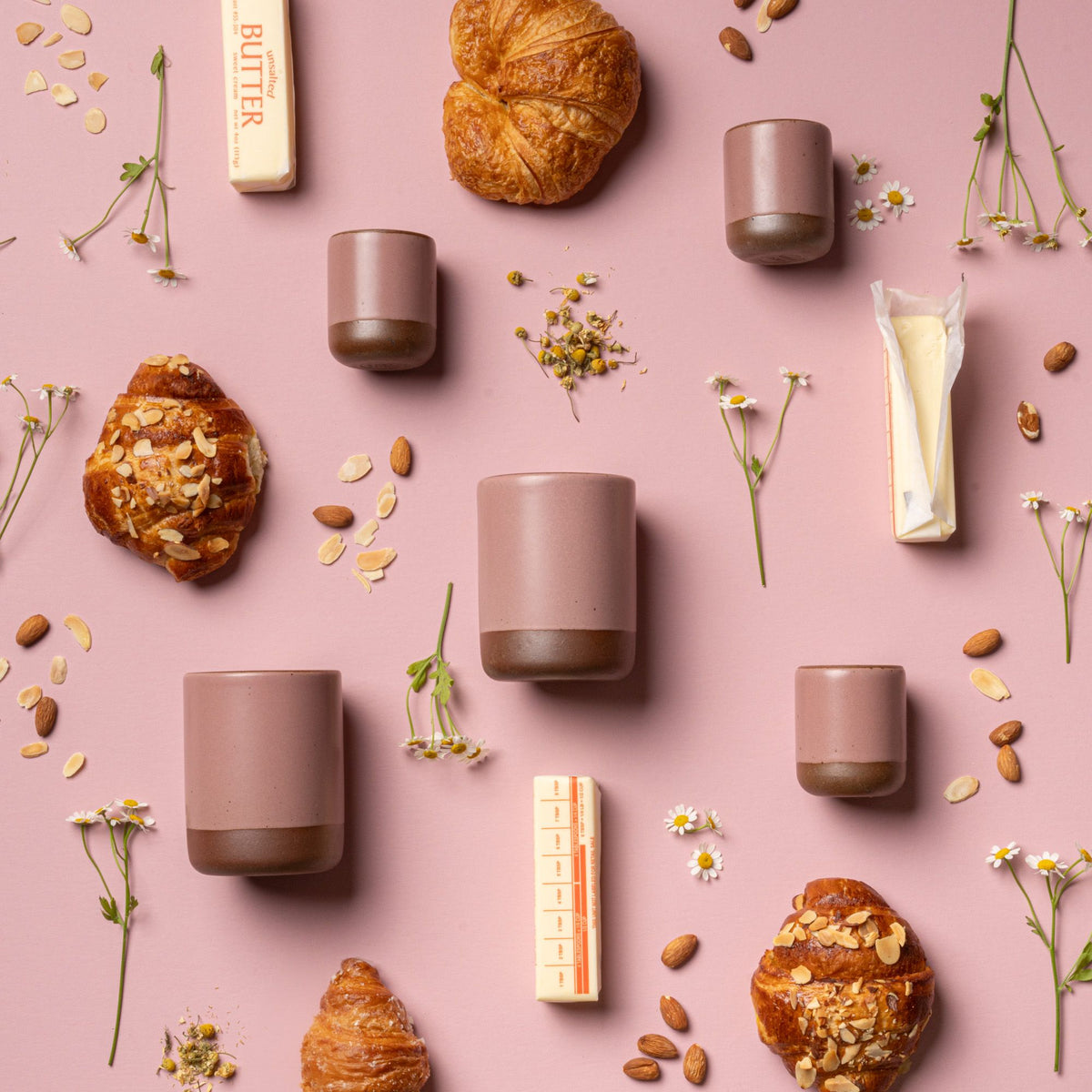 An artful layout of The Candle in Almond Flower - small and large ceramic candles in a cylindrical vessel in a soft mauve pink color, surrounded by almonds butter, and tiny flowers.