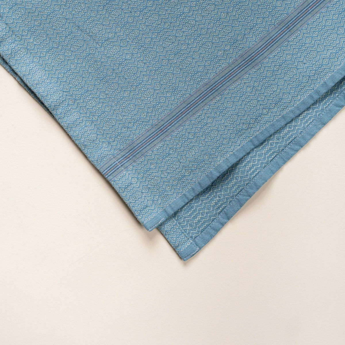 Corner of a bue woven kitchen towel with a subtle textured pattern and slim striped accent, folded neatly on a clean neutral background.