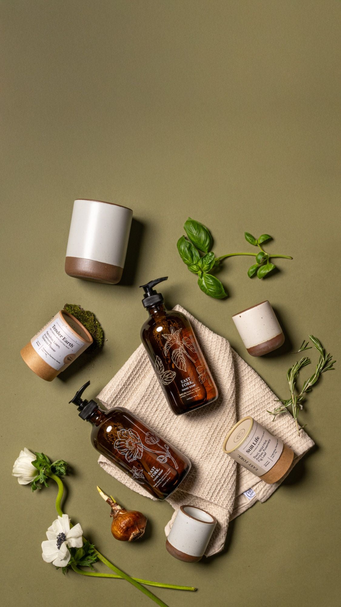 Flat lay of amber soap bottles on a folded towel, ceramic cups, candles, flowers, herbs, and wood sticks on a green background.