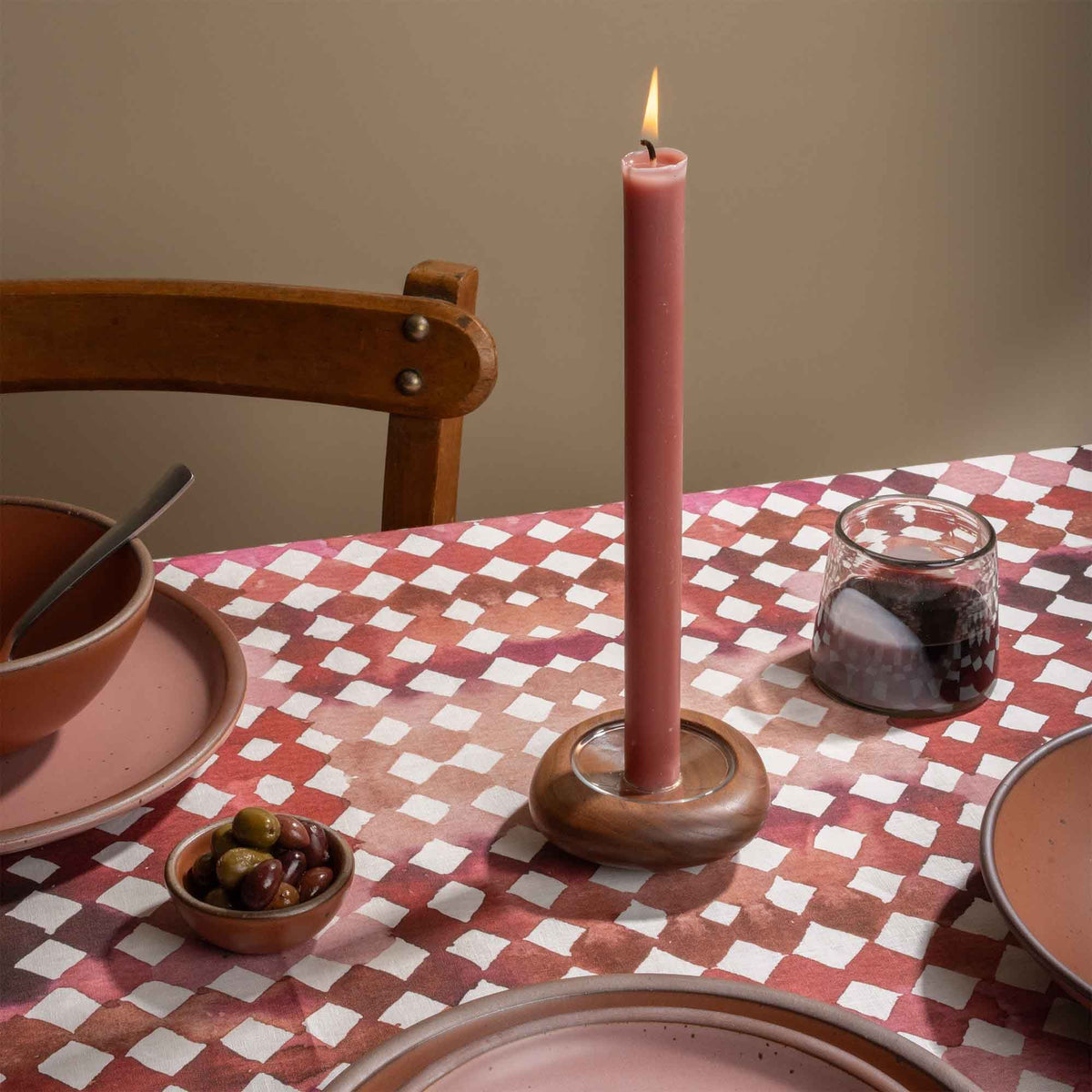 Lit pink taper candle in a wooden holder on a red and pink watercolor diamond tablecloth with tableware and olives.