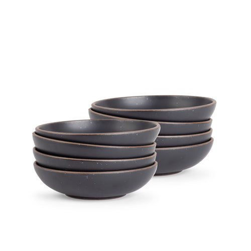 A stack of 8 dinner-sized shallow ceramic bowls in a graphite black color featuring iron speckles and an unglazed rim