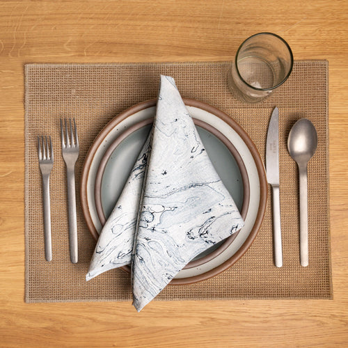 Place setting with stacked plates, blue marbled napkin, silverware, and glass on a woven tan placemat.