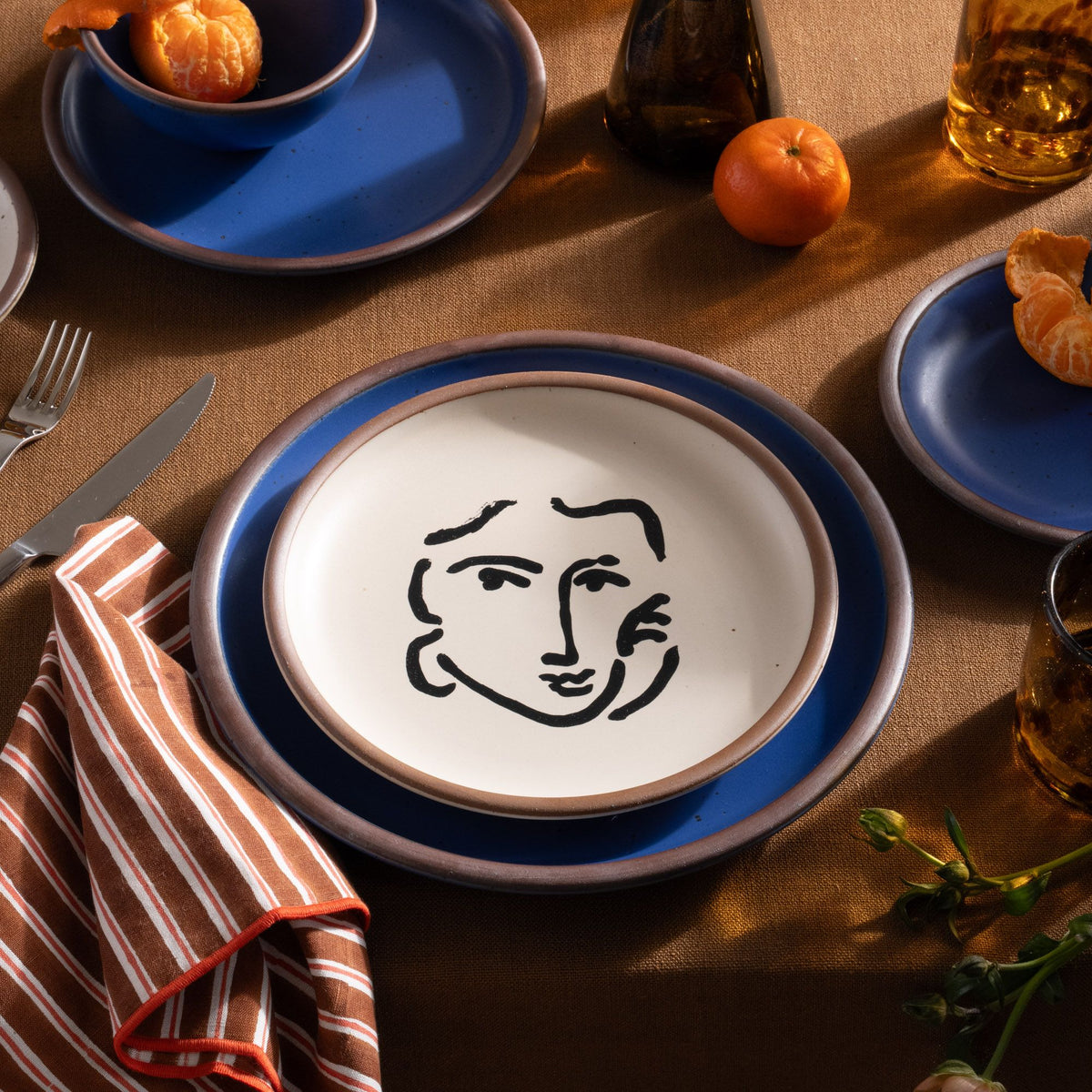 A ceramic plate featuring a decal of the artwork, Nadia au regard sérieux, by Henri Matisse. A minimalist black ink decal of a woman's smiling face in an abstract, expressive style centered on an off-white ceramic plate with an unglazed rim, styled with a blue plate and other dishes on a table with a striped napkin and fruit.