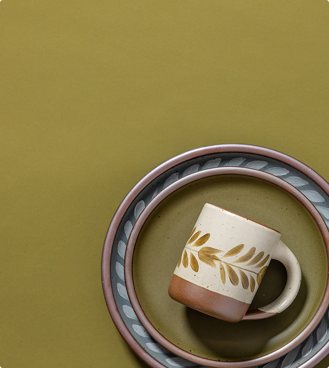 A ceramic mug with a leaf pattern on a cream and brown body, styled on a matching patterned plate against a solid olive green background.
