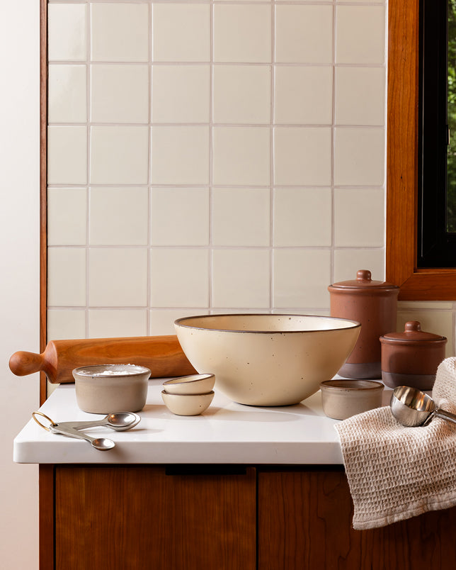 A kitchen counter filled with ceramic bowls, ramekins and jars along with baking tools such as rolling pin, measuring spoons and more.