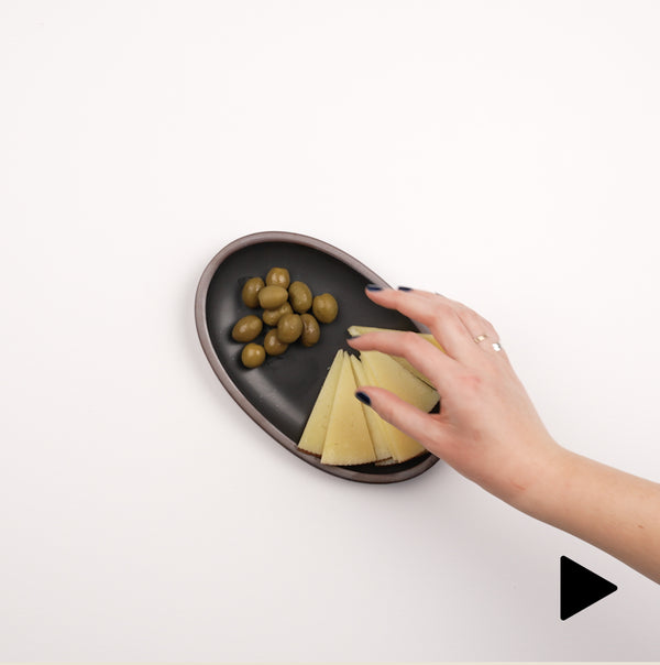 Video of a hand placing a small oval platter in a blue color down filled with olives and cheese slices, and a hand grabbing an olive.