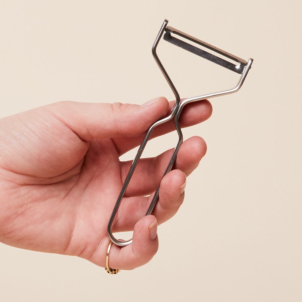 A hand holds a peeler made of metal that has a tapered neck